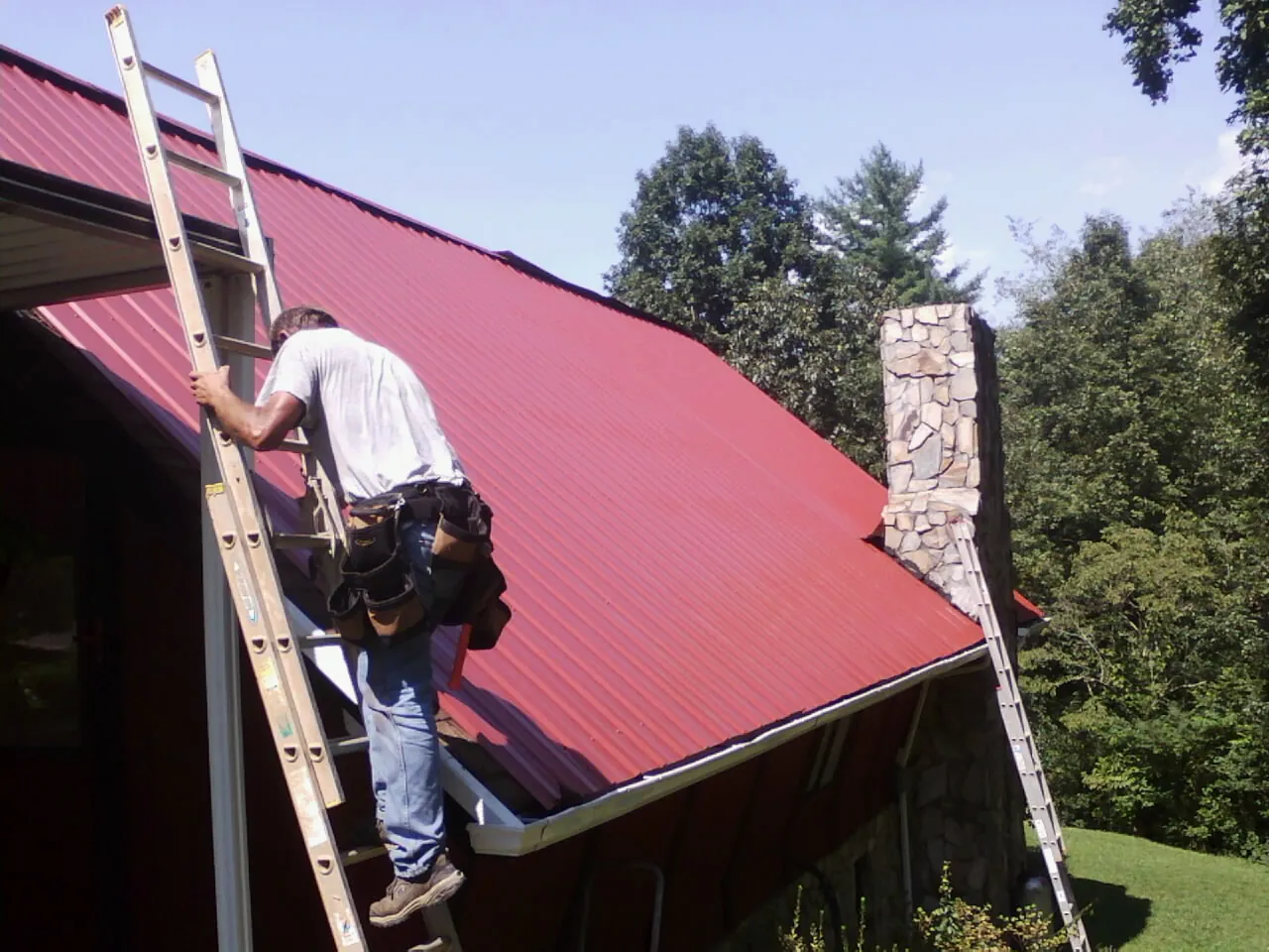 Roofer on a ladder beside a red metal roof and stone chimney — Soffit & Fascia Repair by Roofing Brothers in Stilesville